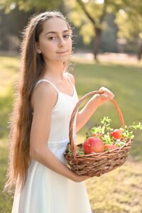 ילדה בשמלת בת מצווה מחזיקה סלסלת רימונים ופירות, עומדת בגינה ירוקה.