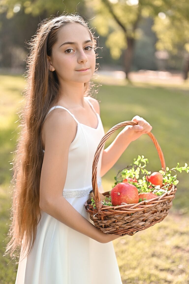 ילדה בשמלת בת מצווה מחזיקה סלסלת רימונים ופירות, עומדת בגינה ירוקה.