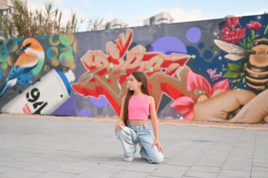 ילדה עם חולצת ורודה וג'ינס מול קיר גרפיטי צבעוני עם ציורי ציפורים ודבורים
