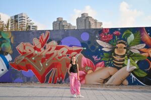 ילדה עם חולצה שחורה ומכנסיים ורודים עומדת לפני קיר גרפיטי עם ציורים של דבורים וצמחים.